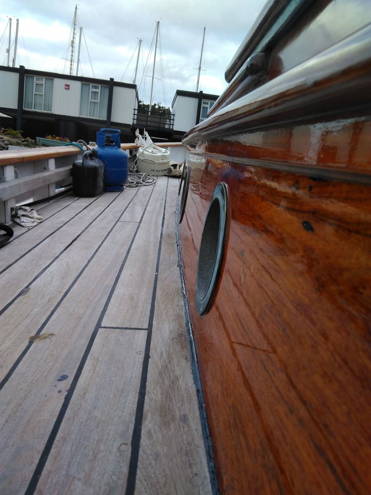 Low view along a laid teak deck beside a deeply varnished cabin side with bronze portlights