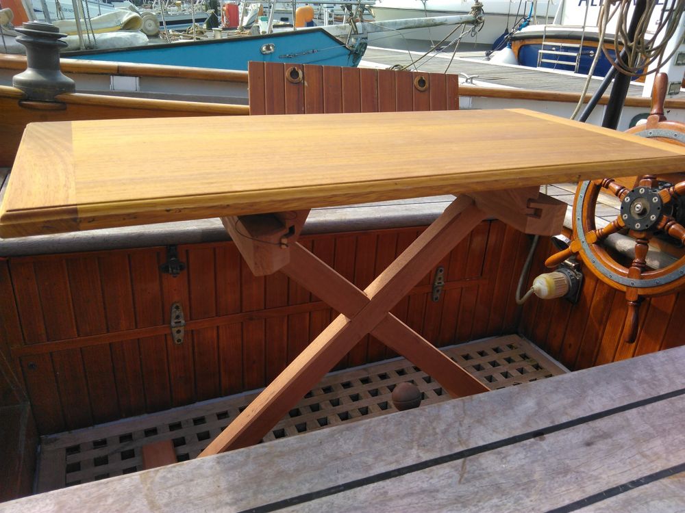 Folding hardwood cockpit table with a framed top and cross-halved X legs, set up in a varnished cockpit