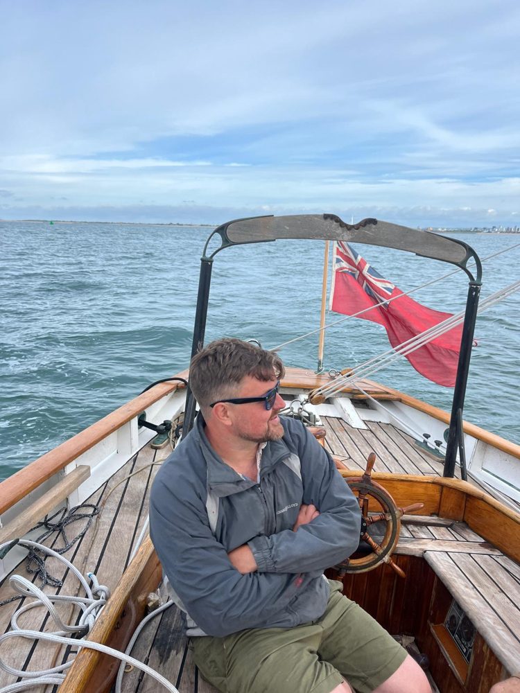 Josh Brookes aboard Rhoda, sitting by the wheel under sail with the red ensign flying astern