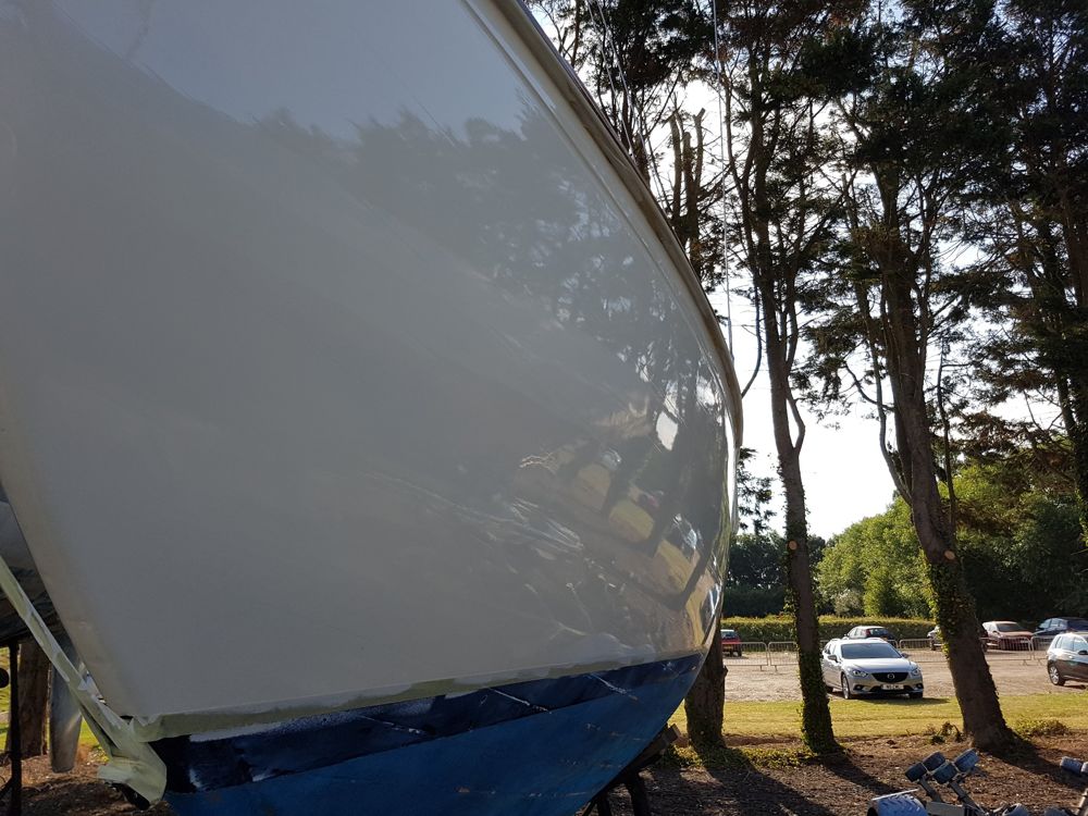 Freshly painted white topsides with a deep gloss reflecting the trees, masking tape still on the boot-top