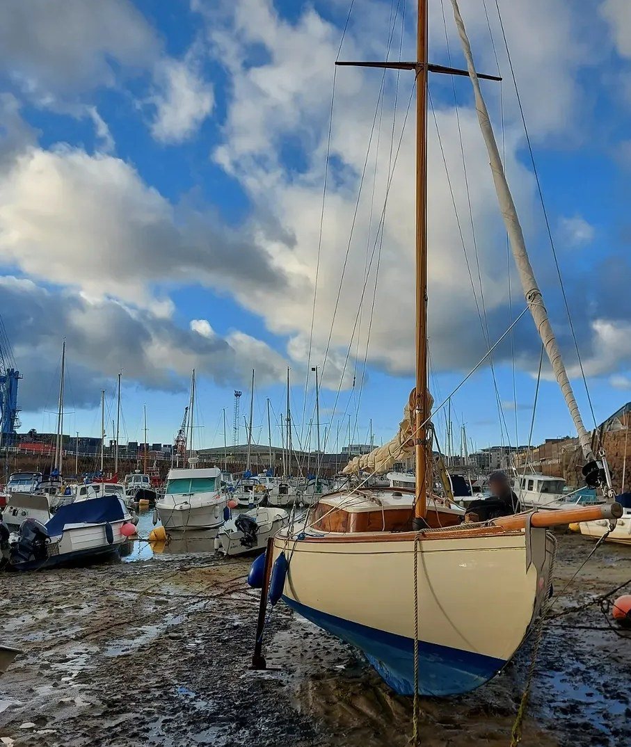 Petrina on legs at low tide in St Helier harbour, her varnished mast and bowsprit against a blue sky