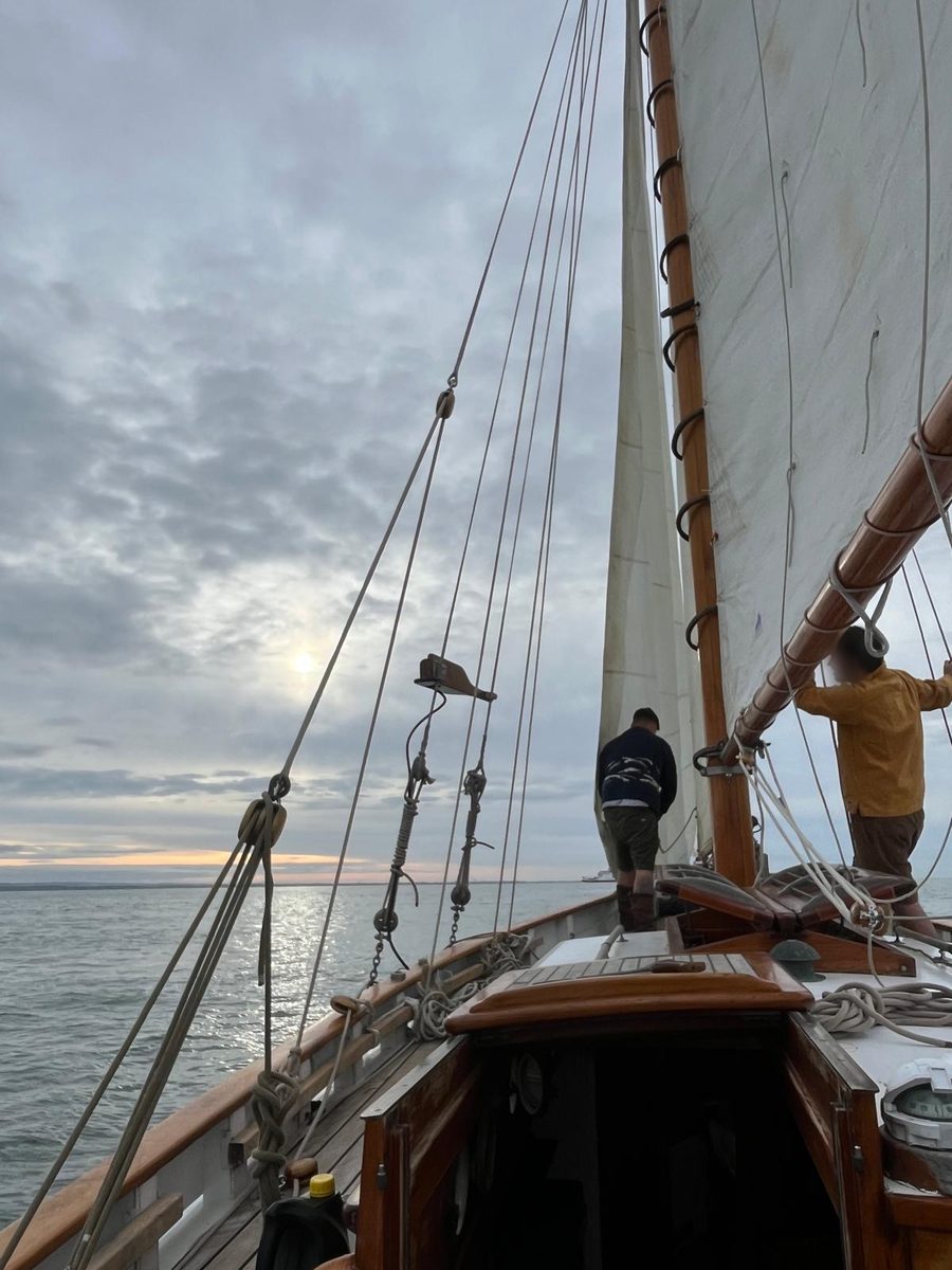 Two crew on Rhoda's foredeck at dusk, the gaff mainsail and wooden mast hoops beside them