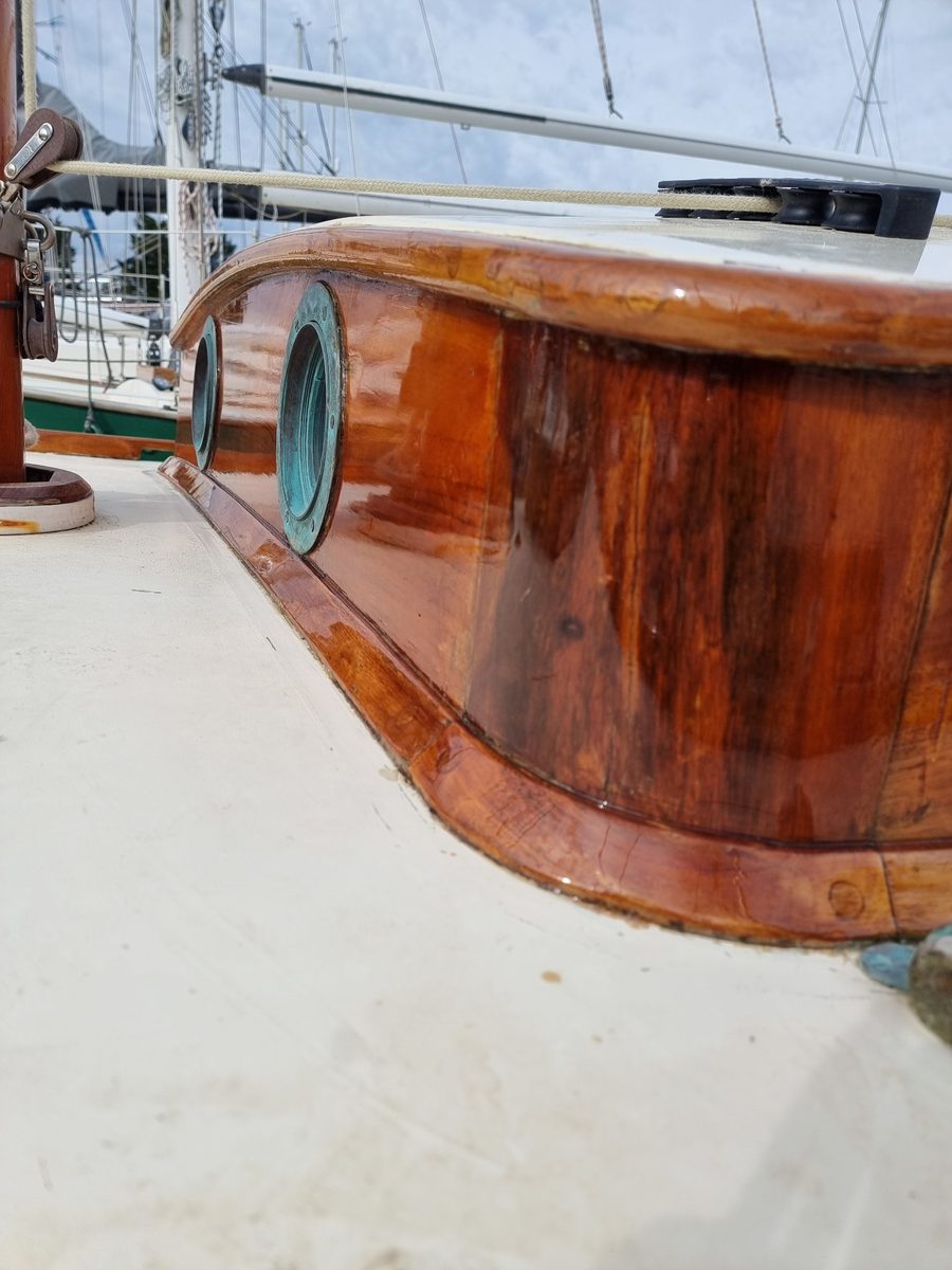 Low view along Roamer's varnished coachroof side and toe rail, with bronze portlights set in the gloss