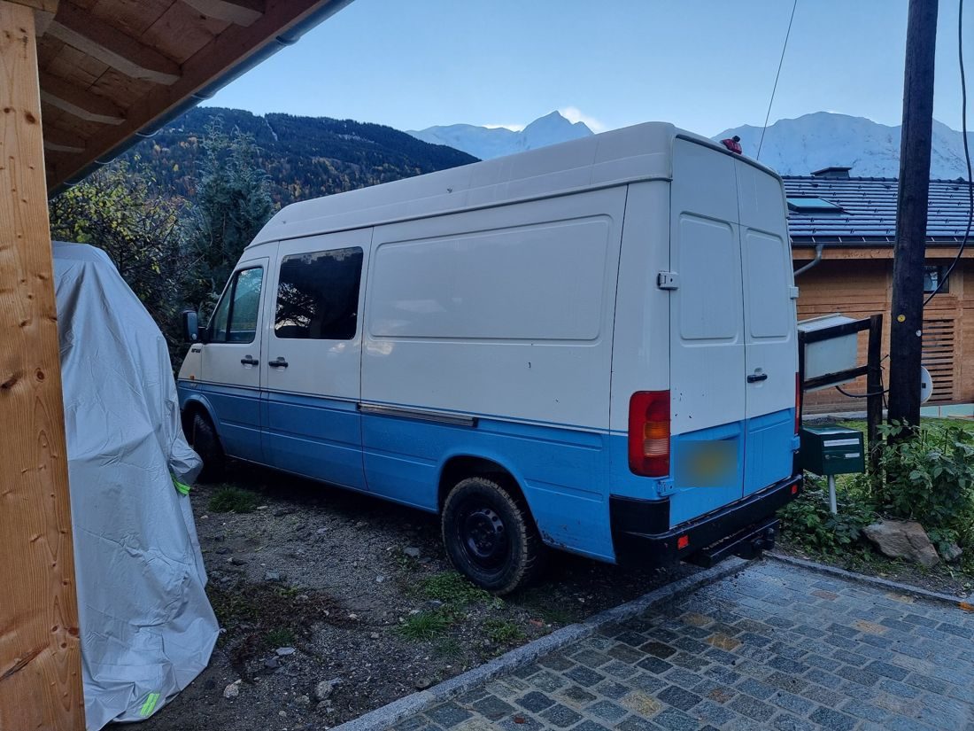 The camper van parked in the Alps, cream above and blue below, with snowy peaks behind
