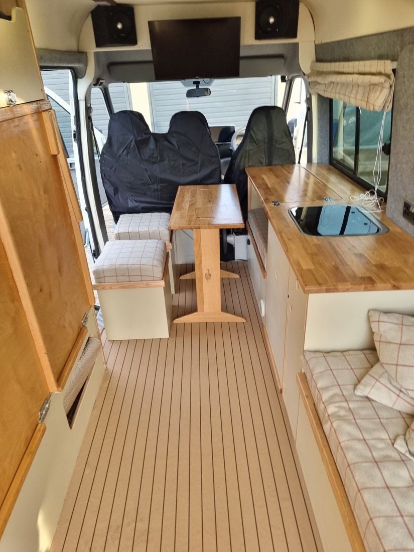 Finished van interior looking forward: oak table, galley worktop with sink, checked cushions and a deck-style floor